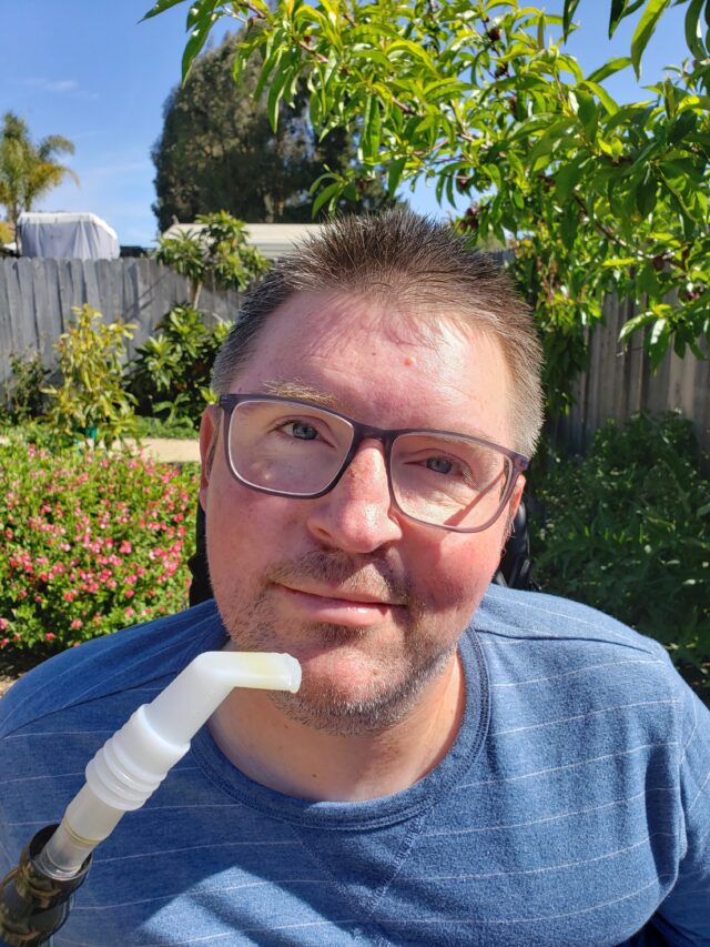 An image of Brian, a white man in a wheelchair with a ventilator mouthpiece. He is outside, with green trees in the background, wearing glasses and a light blue shirt.