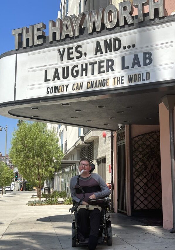 Brian in his wheelchair in a gray and maroon shirt with black pants in front of a marquee for The Hayworth theater with the text "Yes, And… Laughter Lab – Comedy Can Change the World